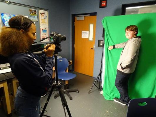 This is a photo of a female student filming a male student who is posing in front of a green screen in a media lab.