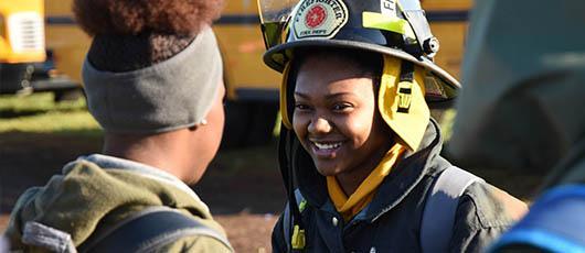 This is a photo of a female student from PSLA at Fowler High School dressed in Fire Rescue gear, smiling at a classmate.