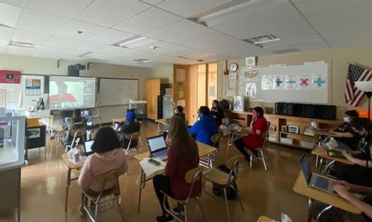 This is a photo from the back of a classroom showing students at their desks watching a screen projection showing a Corcoran alumni who was guest speaking.